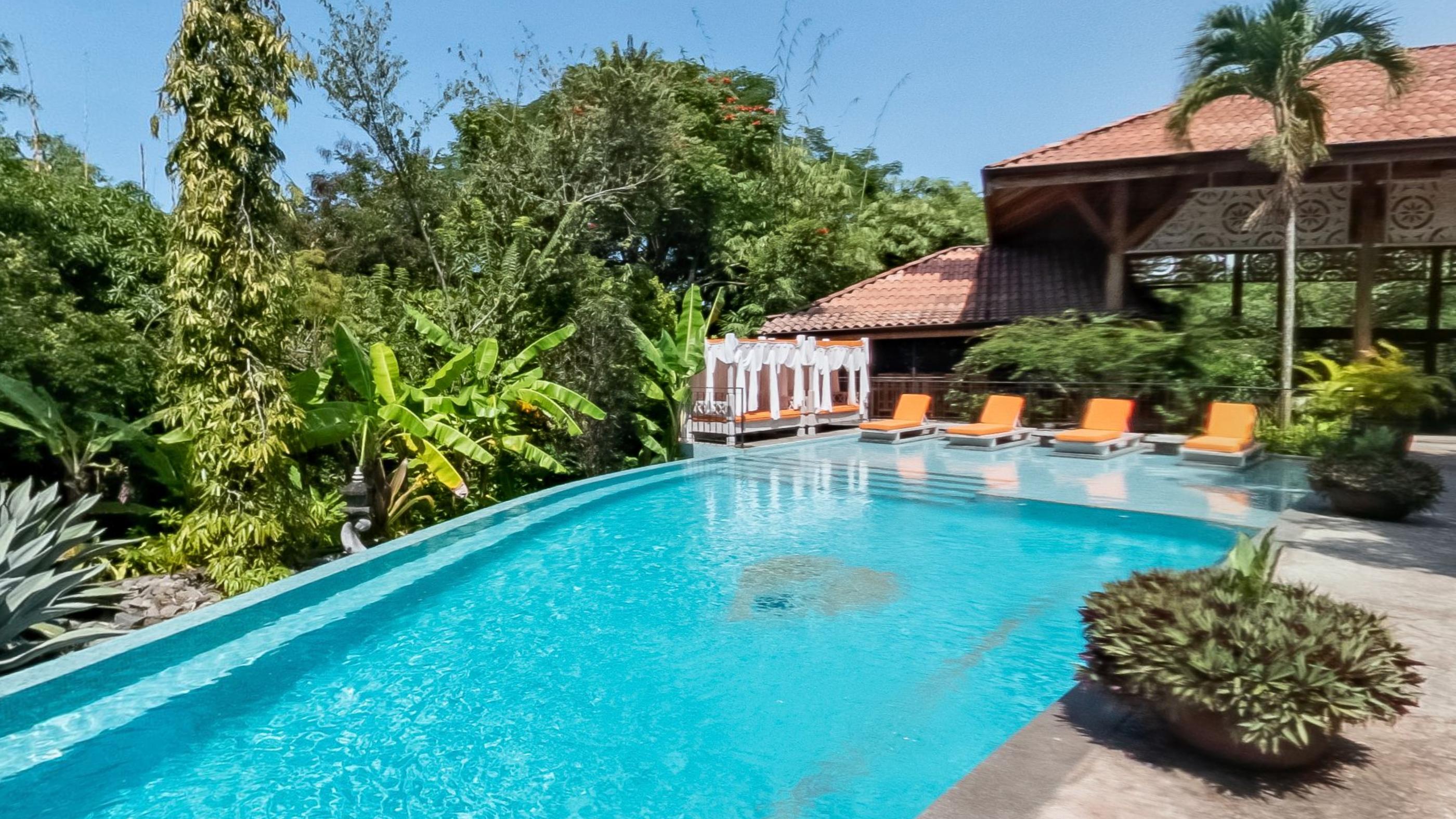 A long turquoise pool with orange loungers along it and a tiled timber pavilion at the far end