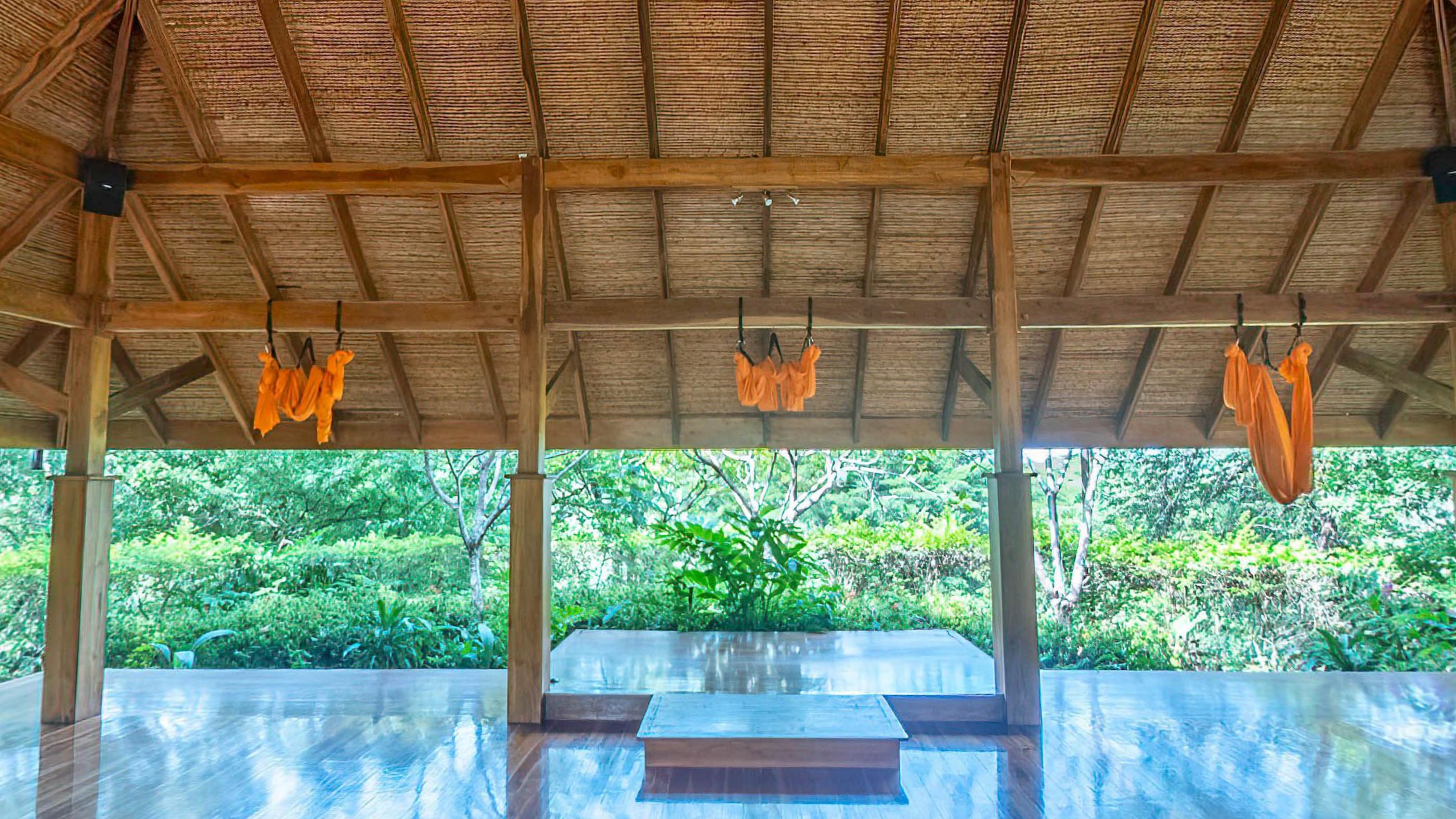An open sided yoga hall with a polished floor, orange aerial hammocks hanging from the beams and jungle on every side