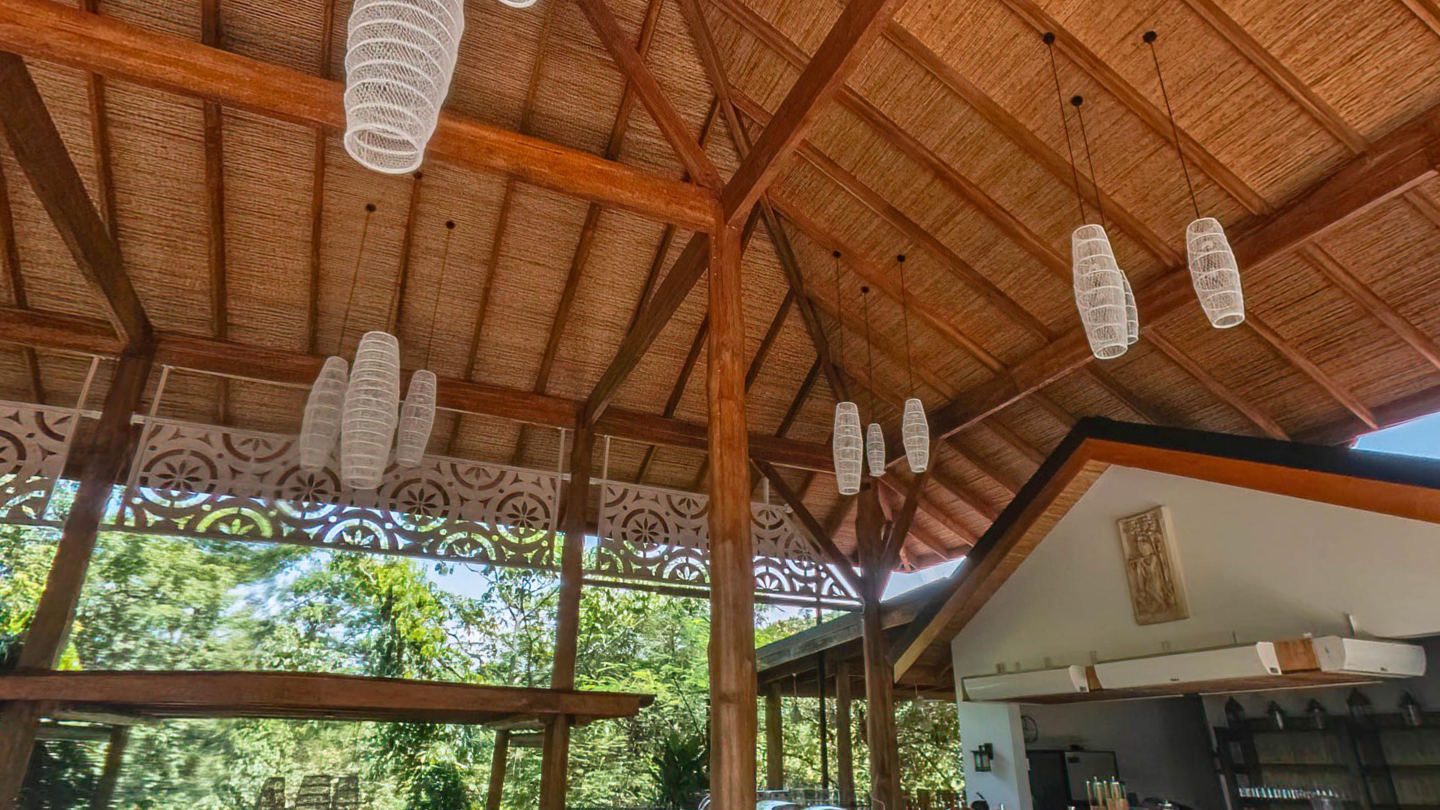A woven bamboo ceiling with timber rafters, carved fretwork panels and pale pendant lanterns, jungle visible under the open eaves