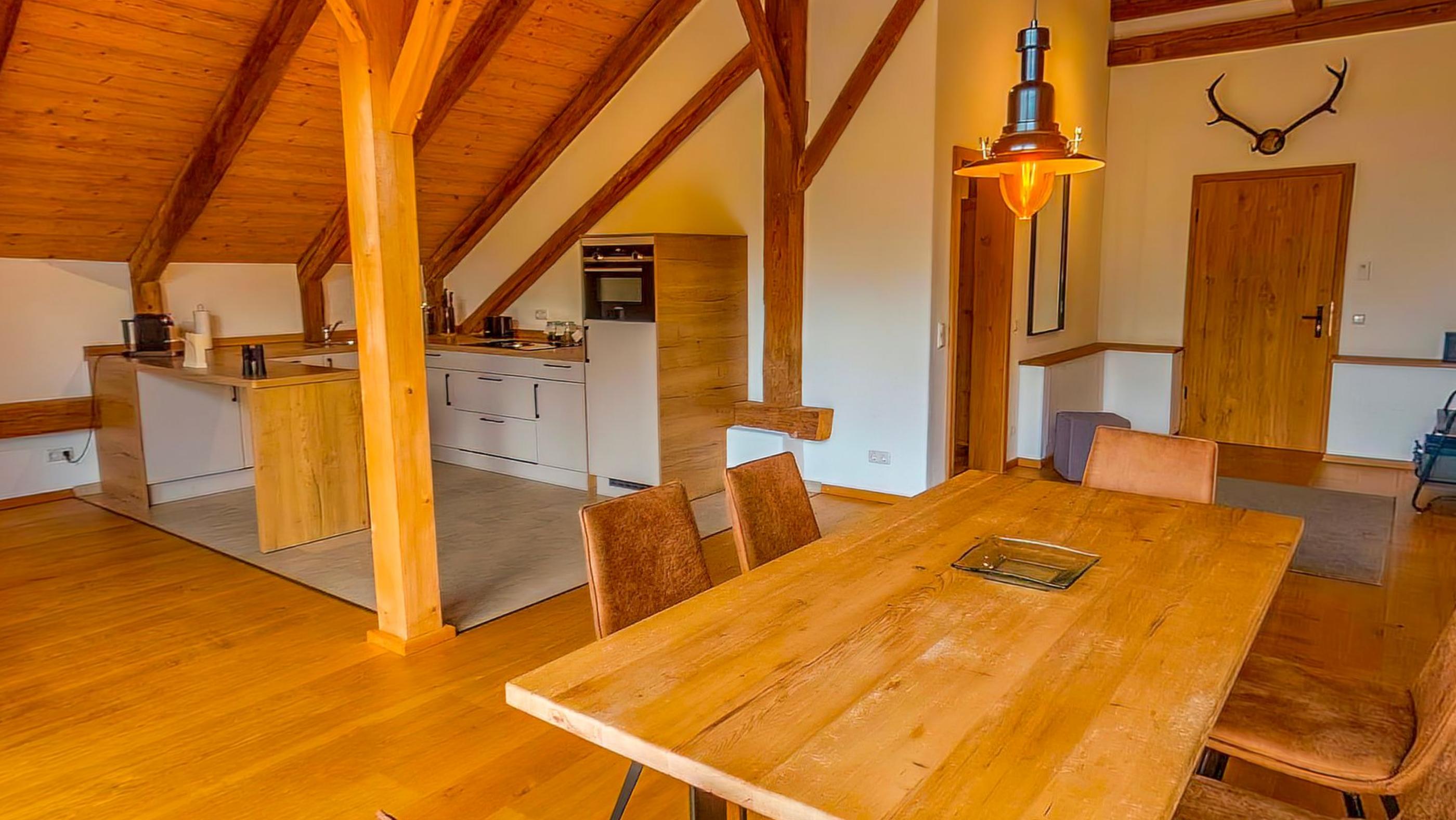 The long timber dining table with the kitchen behind it, antlers on the plaster wall and heavy rafters above