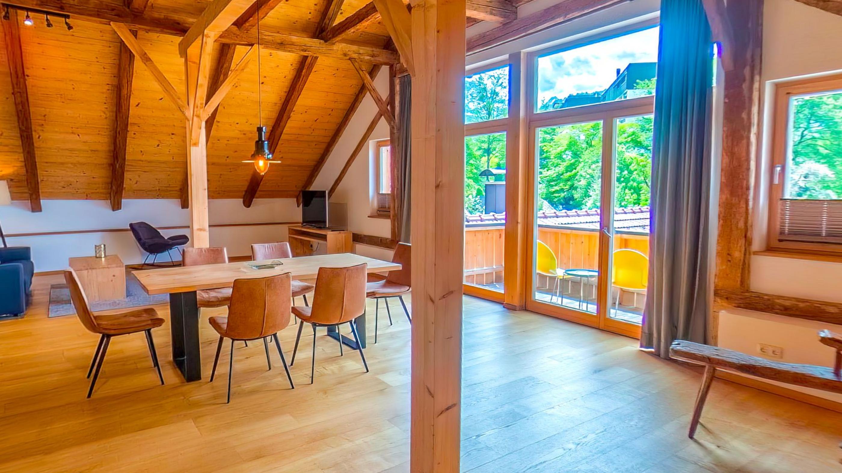 The dining table and chairs in the open attic with a wall of glass onto the balcony and rafters overhead