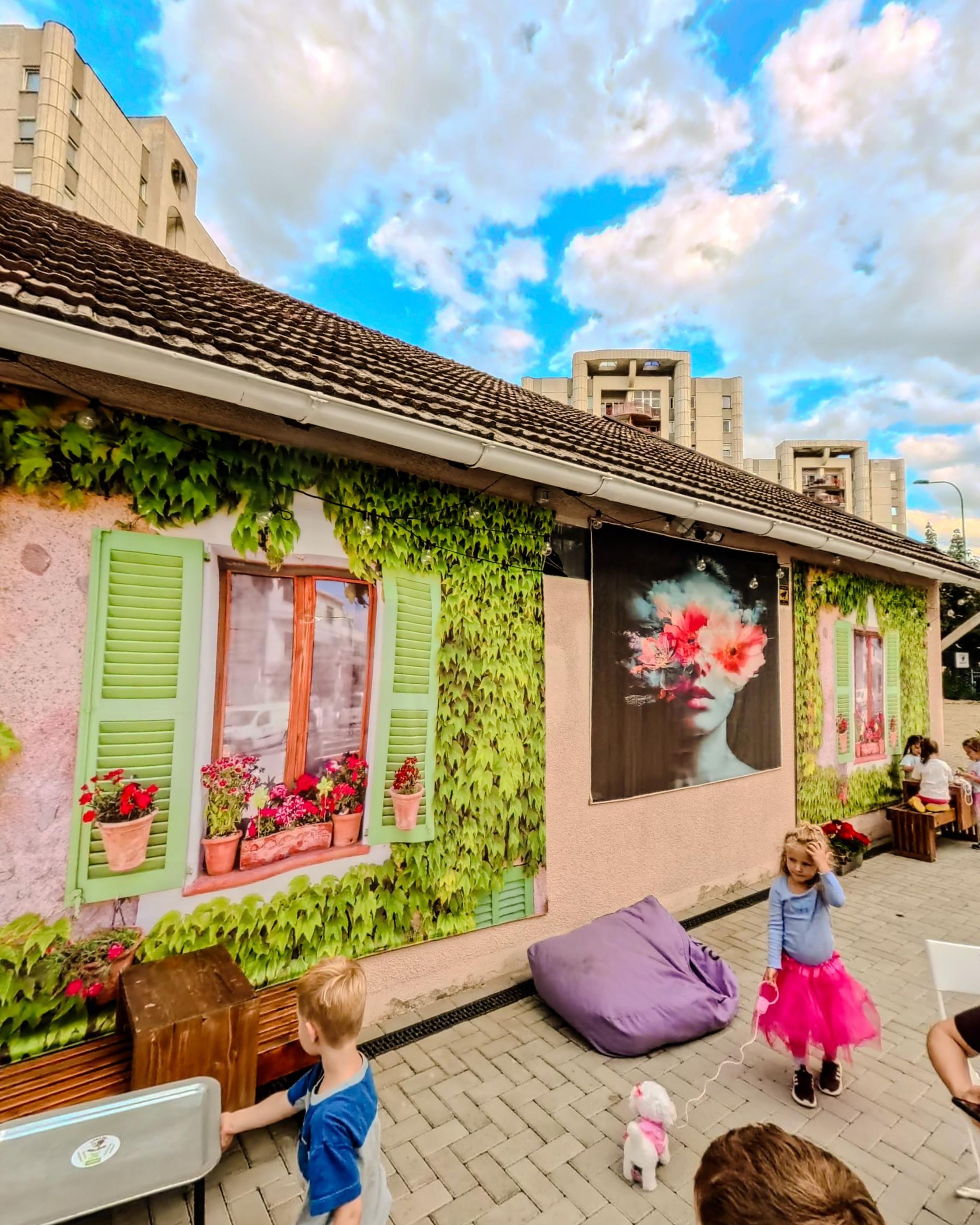 A pink wall under ivy with green shutters, geraniums and a painted mural, children playing in front