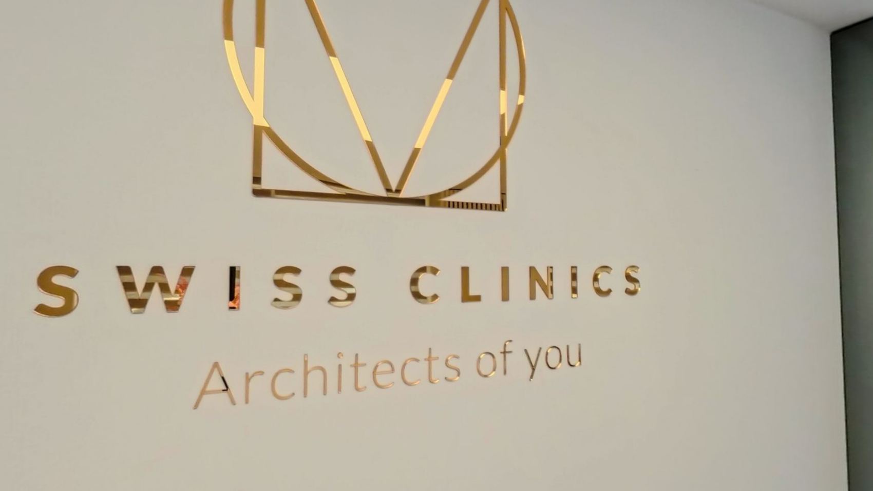 The gold Swiss Clinics wordmark and the words Architects of you, on a white wall