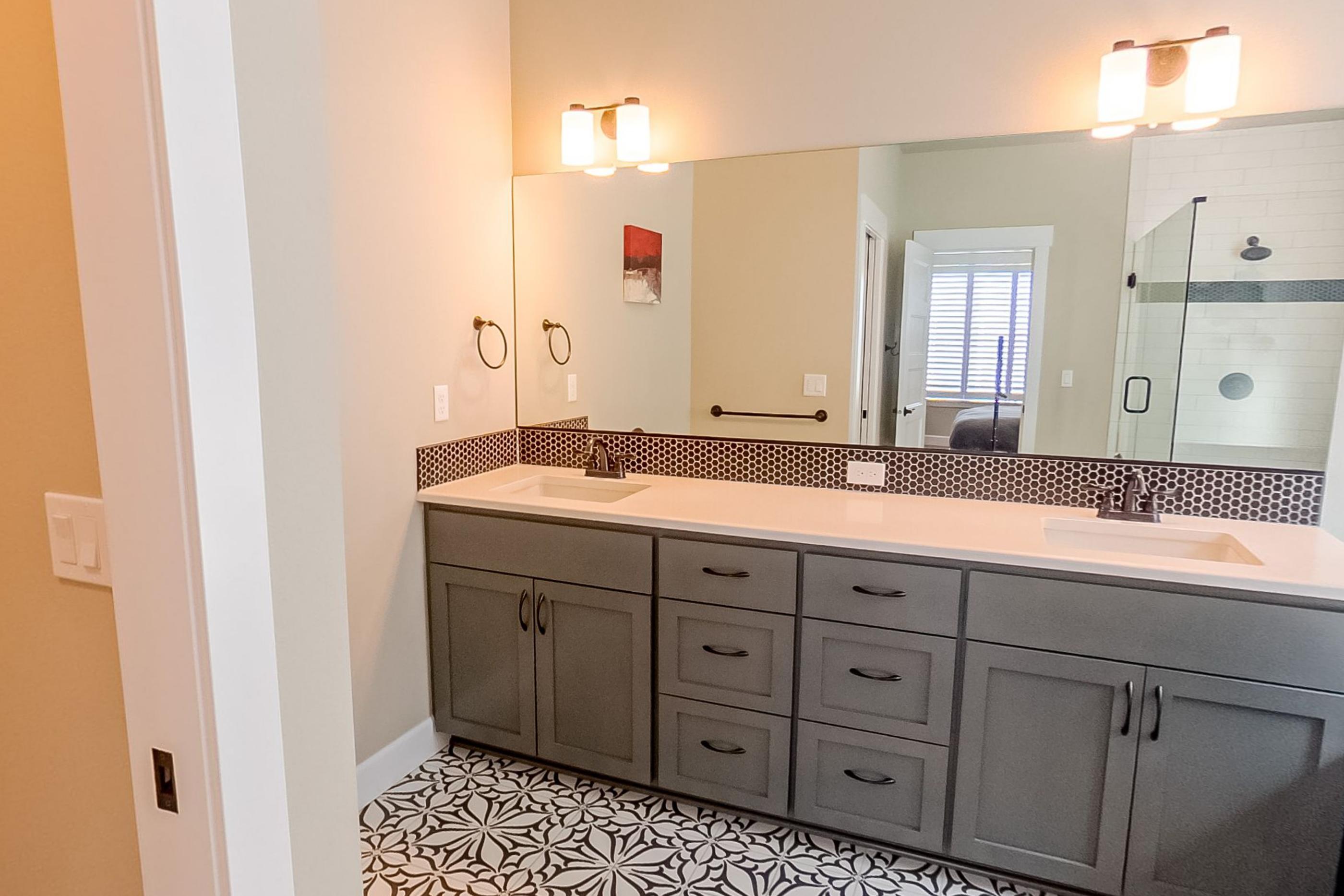 A bathroom with twin basins, a large mirror, patterned floor tile and a glass shower