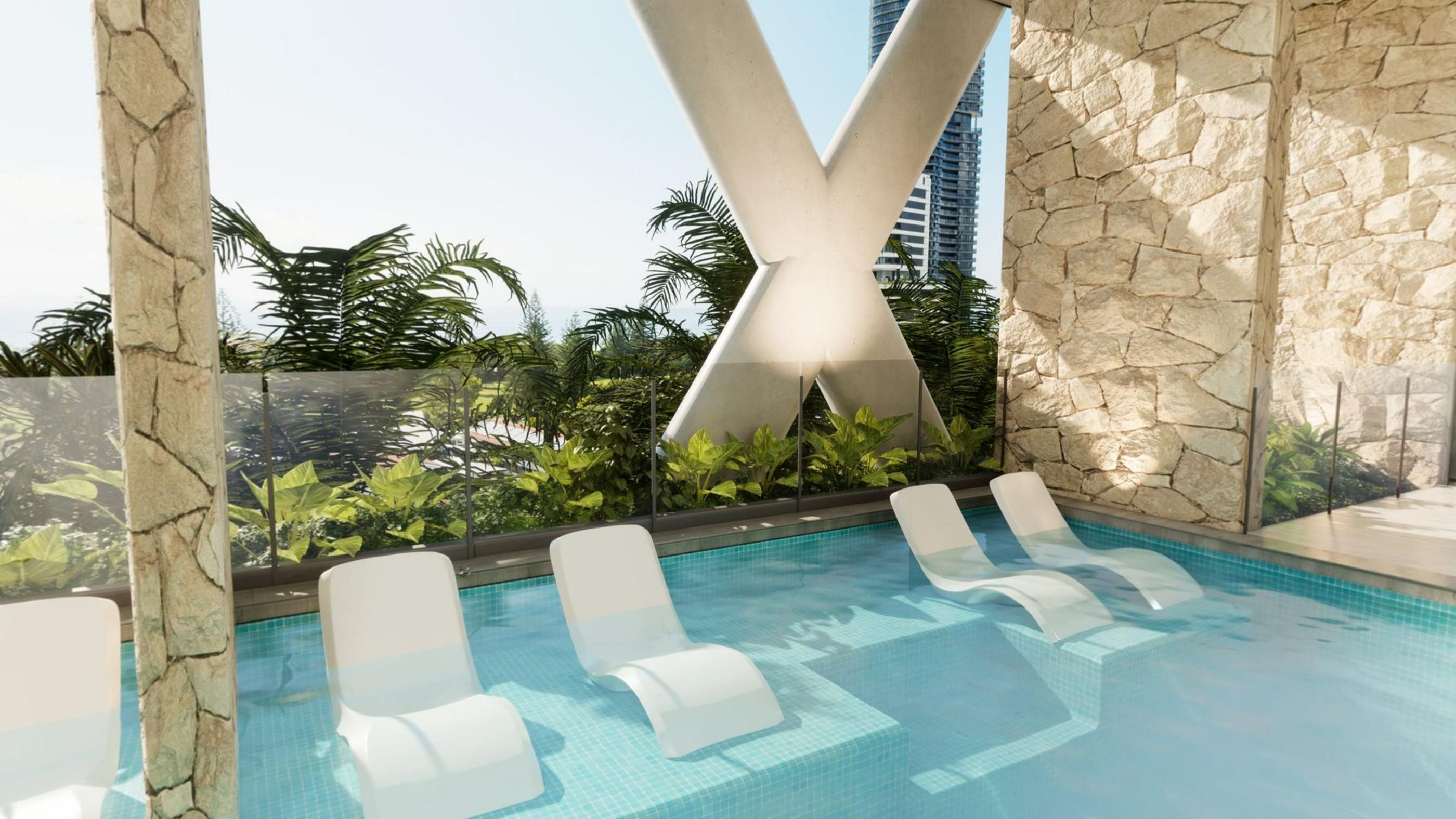 A rectangular pool with white loungers, a stone wall and palms on a podium deck