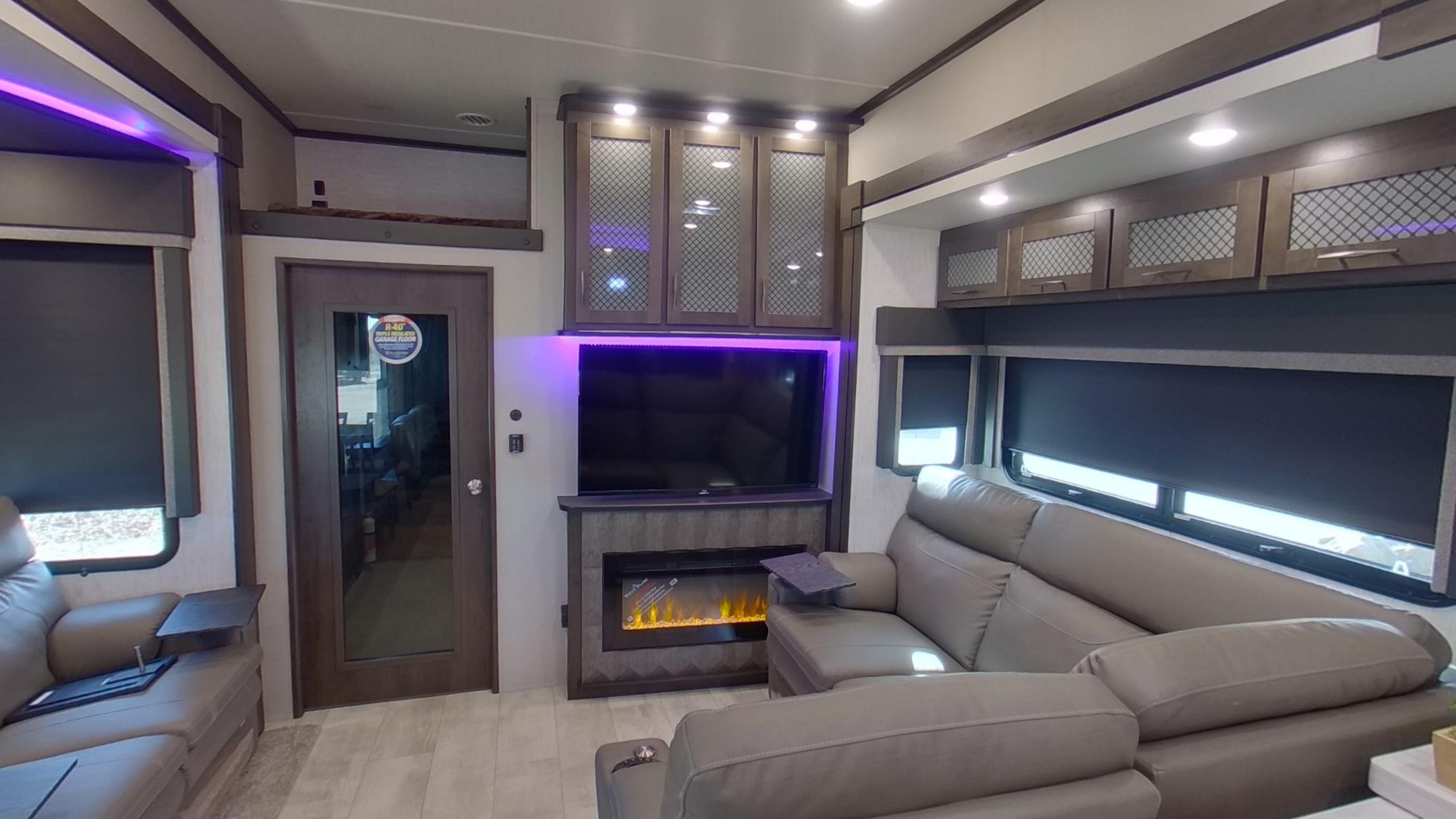 The living area of the RV with a corner sofa, an electric fireplace under a wall mounted television and violet cove lighting